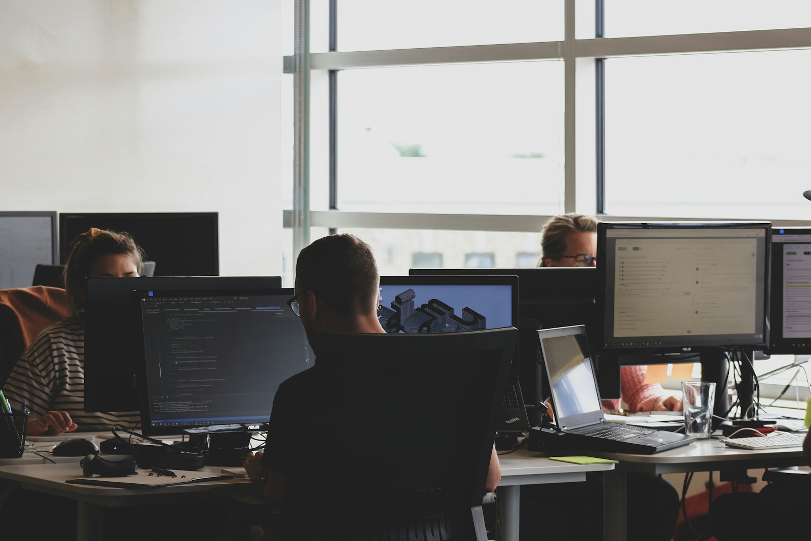 People work at multiple computer monitors showing code and a digital model in an office.