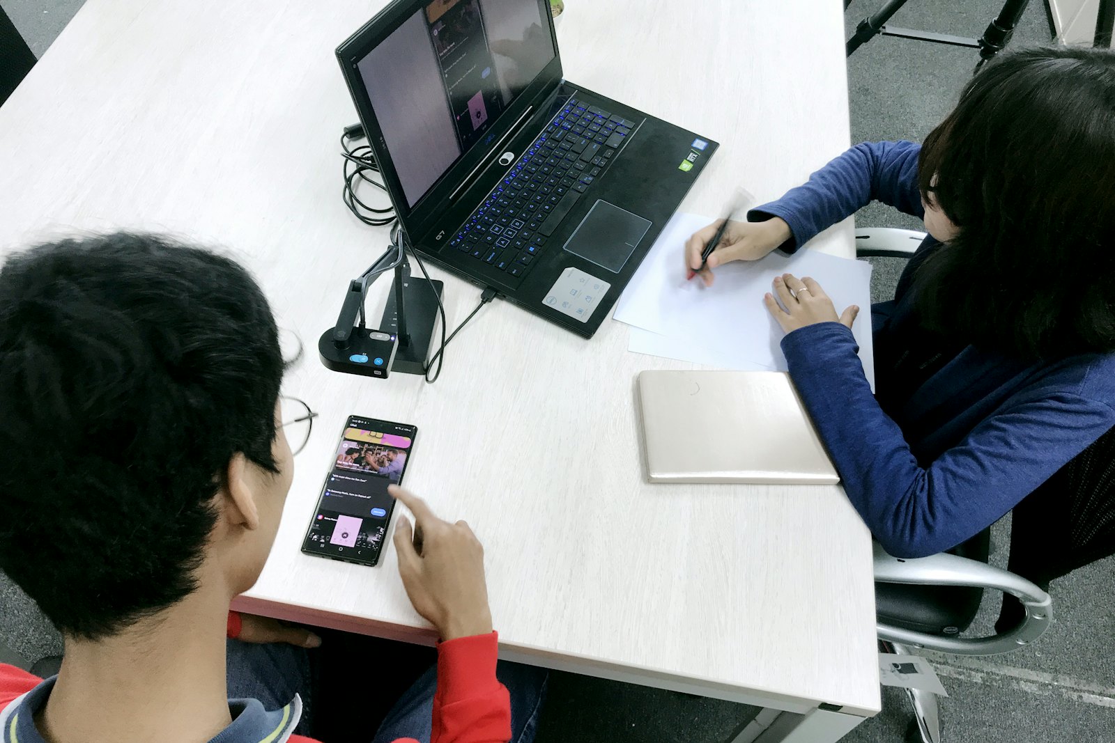 One person uses a phone while another takes notes beside a laptop during a usability session.